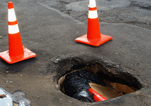 Cave-in on a city street showing a deep hole with jagged edges. Trash bags and a traffic cone are inside the hole.