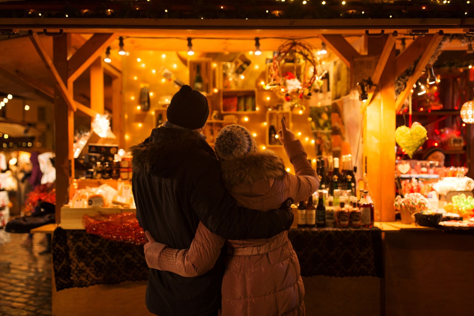 happy senior couple hugging at christmas market souvenir shop stall in evening where woman points at merchandise